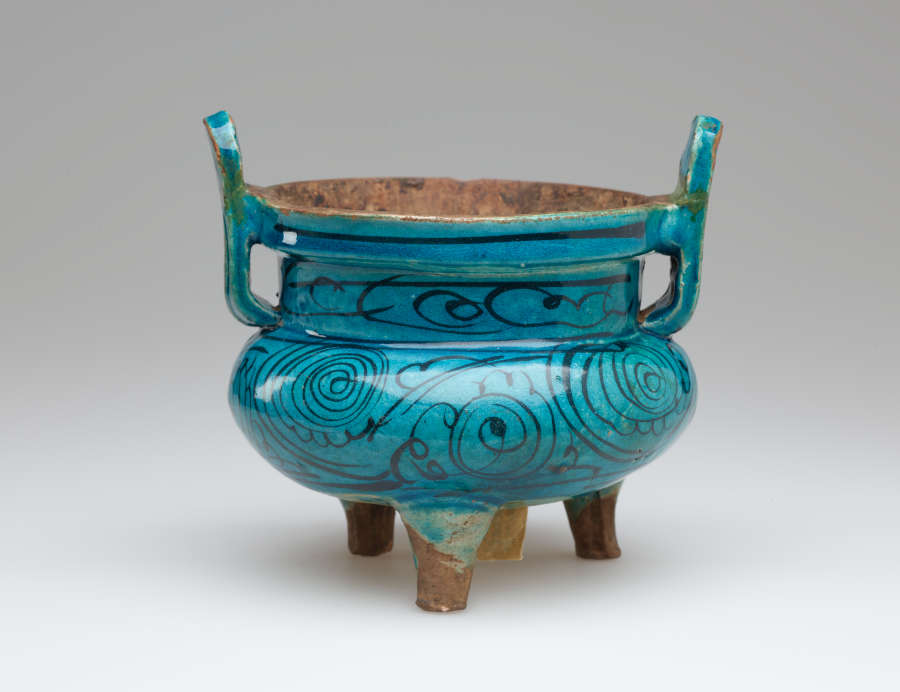 Round, blue glazed vessel with dark thin swirling patterns on body. Two thin handles flare upward. Interior and four feet are brown, unglazed clay.