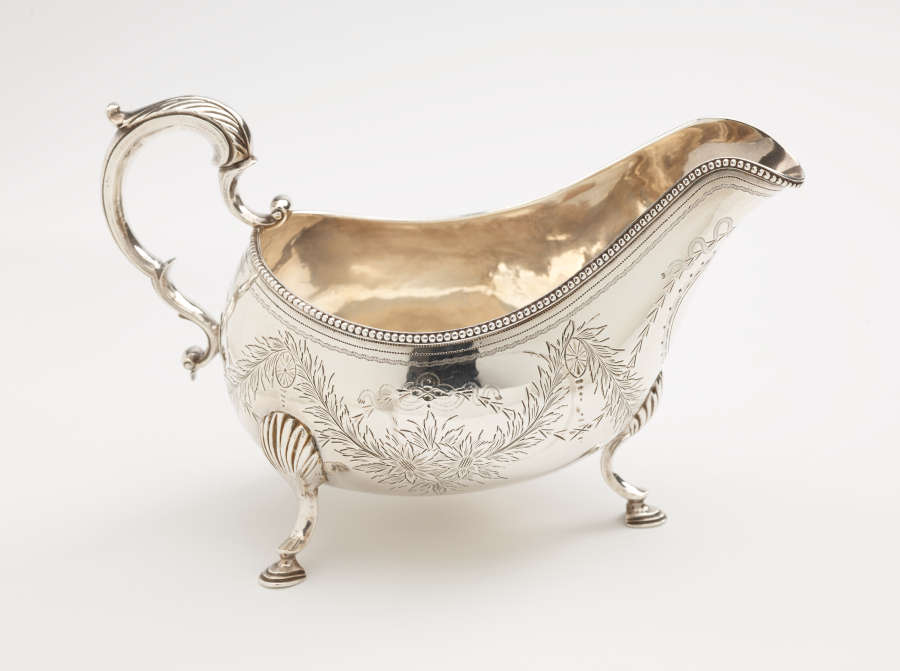 A silver sauce boat with a sculptural handle, high lip for pouring, and three protruding feet. There are floral engravings along the body of the vessel.