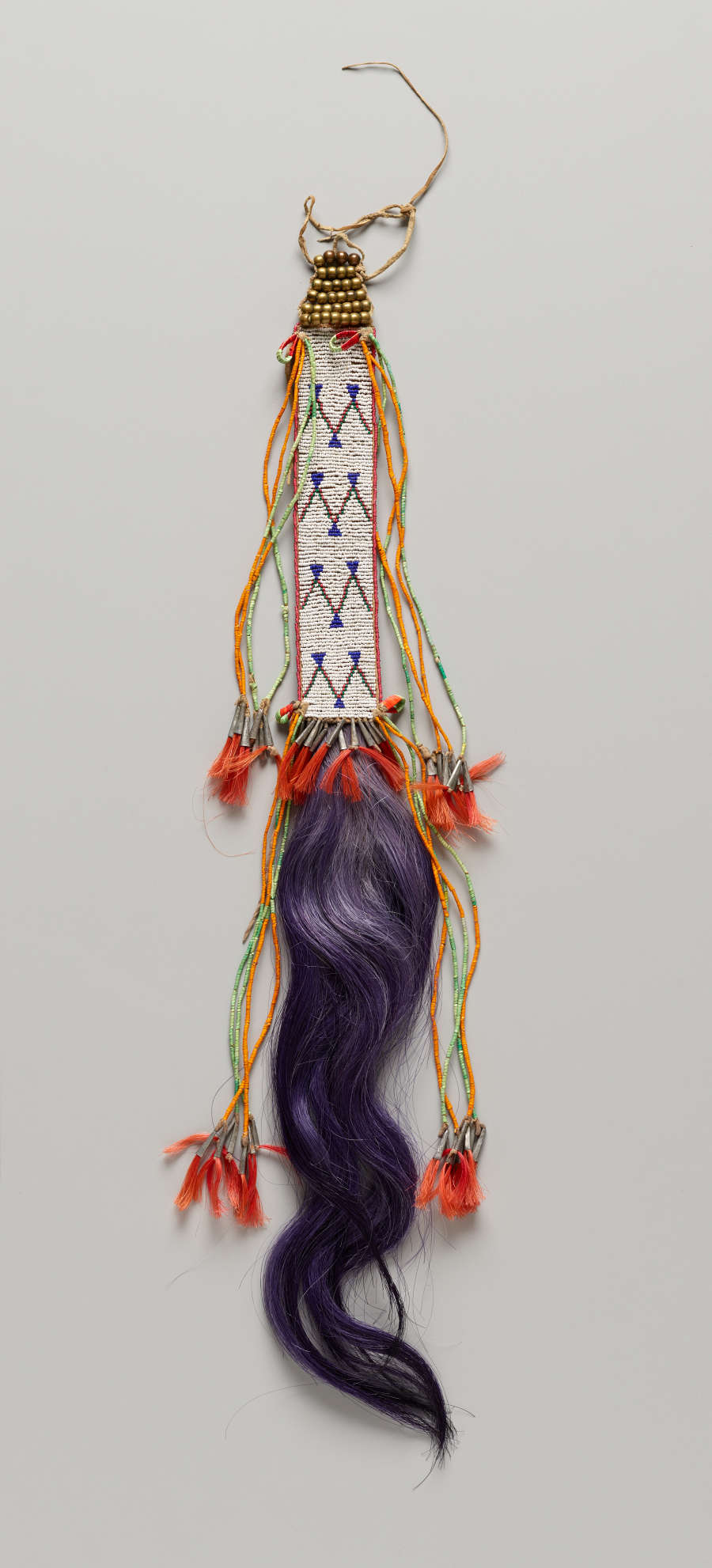 Long vertically beaded rectangle with bright colorful tassels and long wavy purplish hair coming from the bottom. The tassels are orange and connect to green, red, and yellow threads.