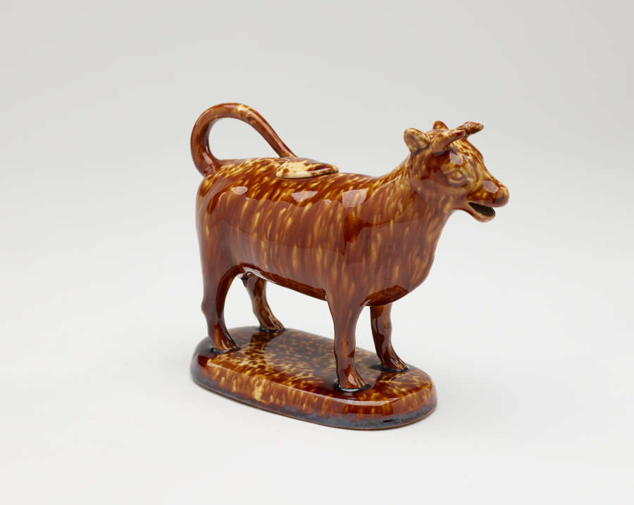  A brown sculptural creamer in the shape of a cow with horns. There is a small flat lid in the center of its back.