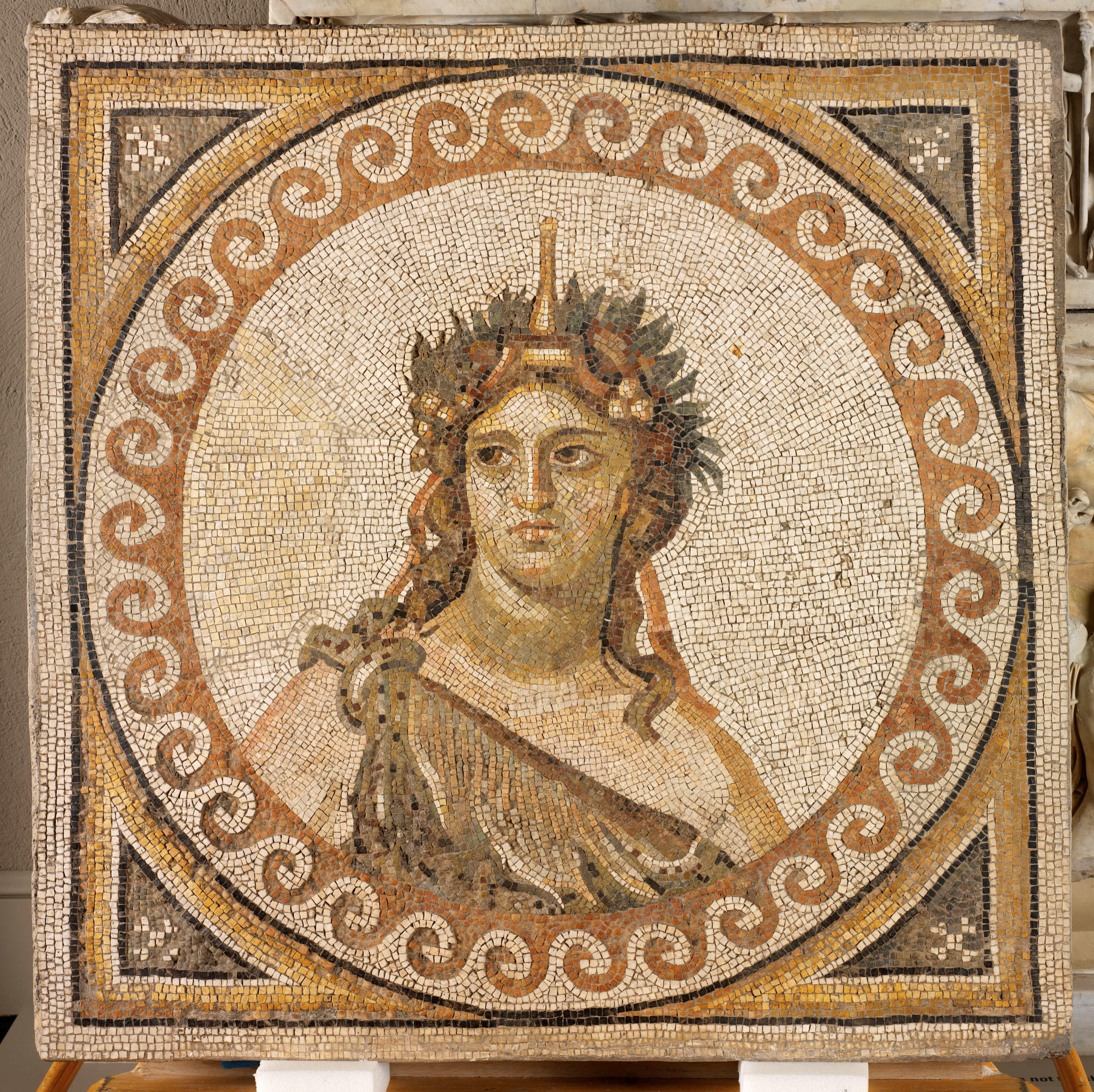 A mosaic depicting a figure with flowing hair, wearing a laurel crown and headpiece and draped robe, enclosed within a circular frame with a repeating wave motif.