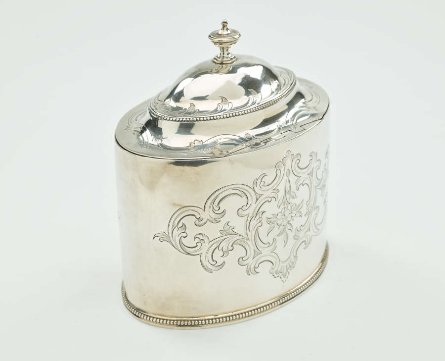 A silver tea caddy with a lid and sculptural finial. There are swirling engravings along the body and lid.