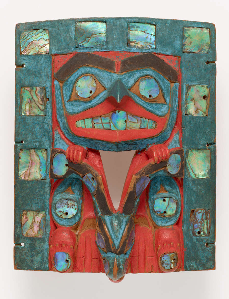 Wooden carving of a face with hands holding onto a deer’s antlers, painted in reds, blues, and ochres. It is framed by a turquoise border patterned with iridescent shell inlays.