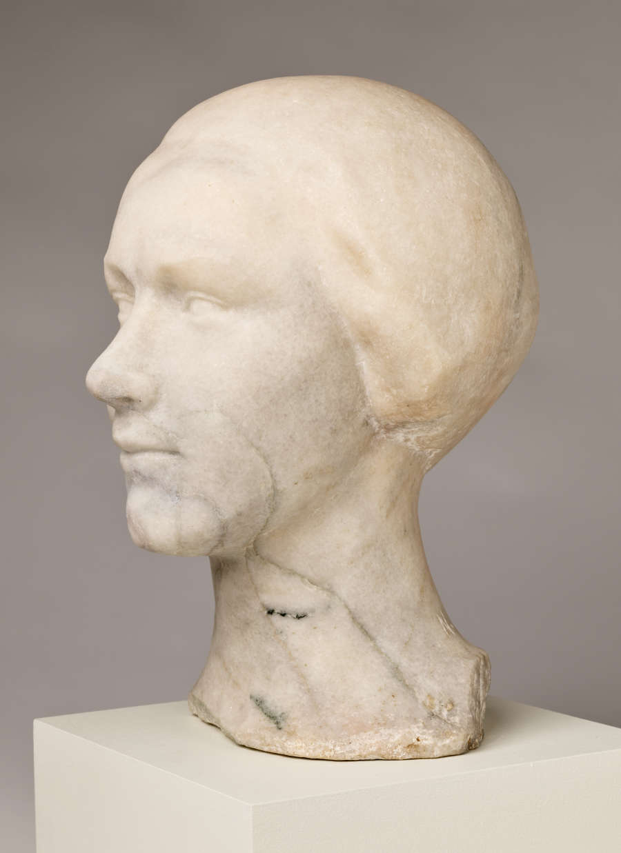 Three quarter front view of a blemished marble sculpture depicting the head of a female figure. The face features a faint smile and delicate details, framed by a short hairstyle.