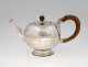 A bulbous silver teapot with a wooden finial and handle connected with decorative silver pieces. There are delicate engraved marks on the body and lid.