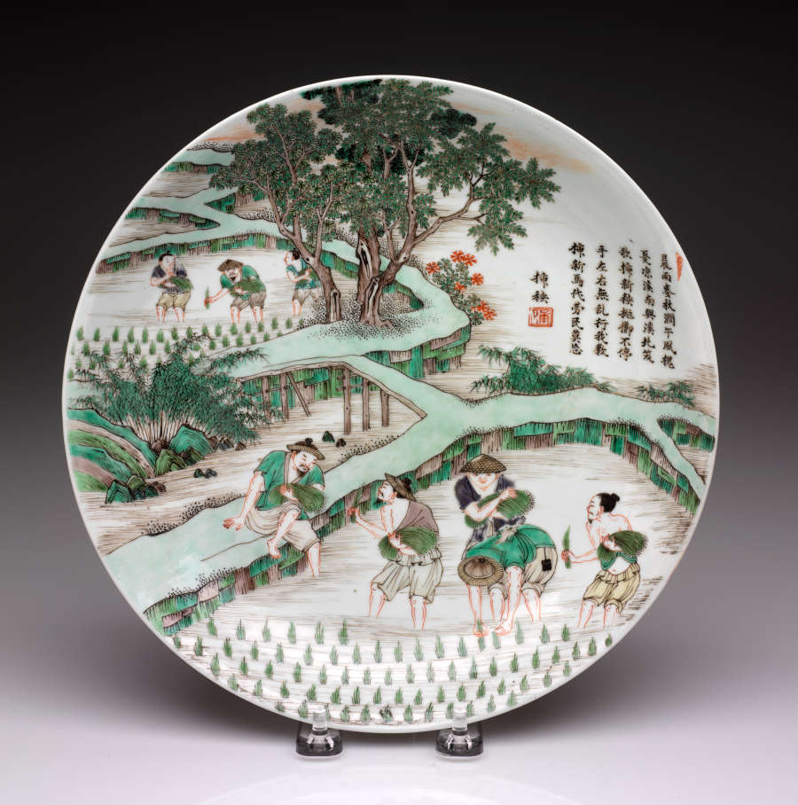 White plate propped on a stand, with vibrant green, red, and yellow illustrations of fars. mers in rice hats tending to paddy fields, surrounded by trees and built walkways.