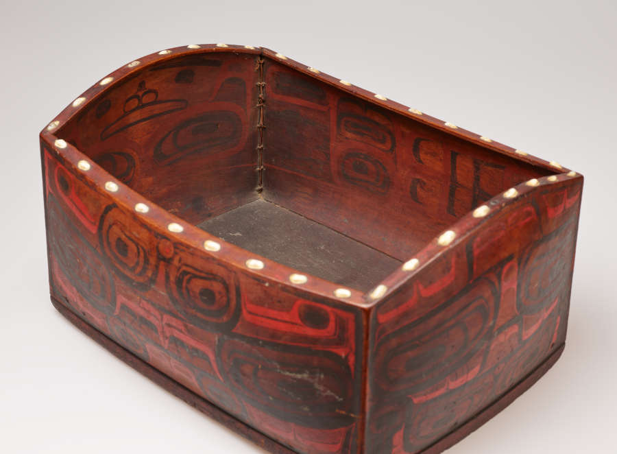 Quarter-view of a bulging rectangular wooden box with faded, red and black, graphic illustrations, some mimicking faces. The box's rim undulates and is decorated with a line of white dots.