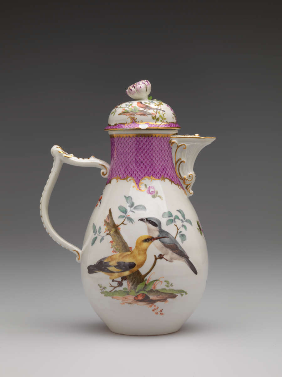 A white coffee pot with gold, pink, and floral decorations. The central decoration has two birds perched on branches.