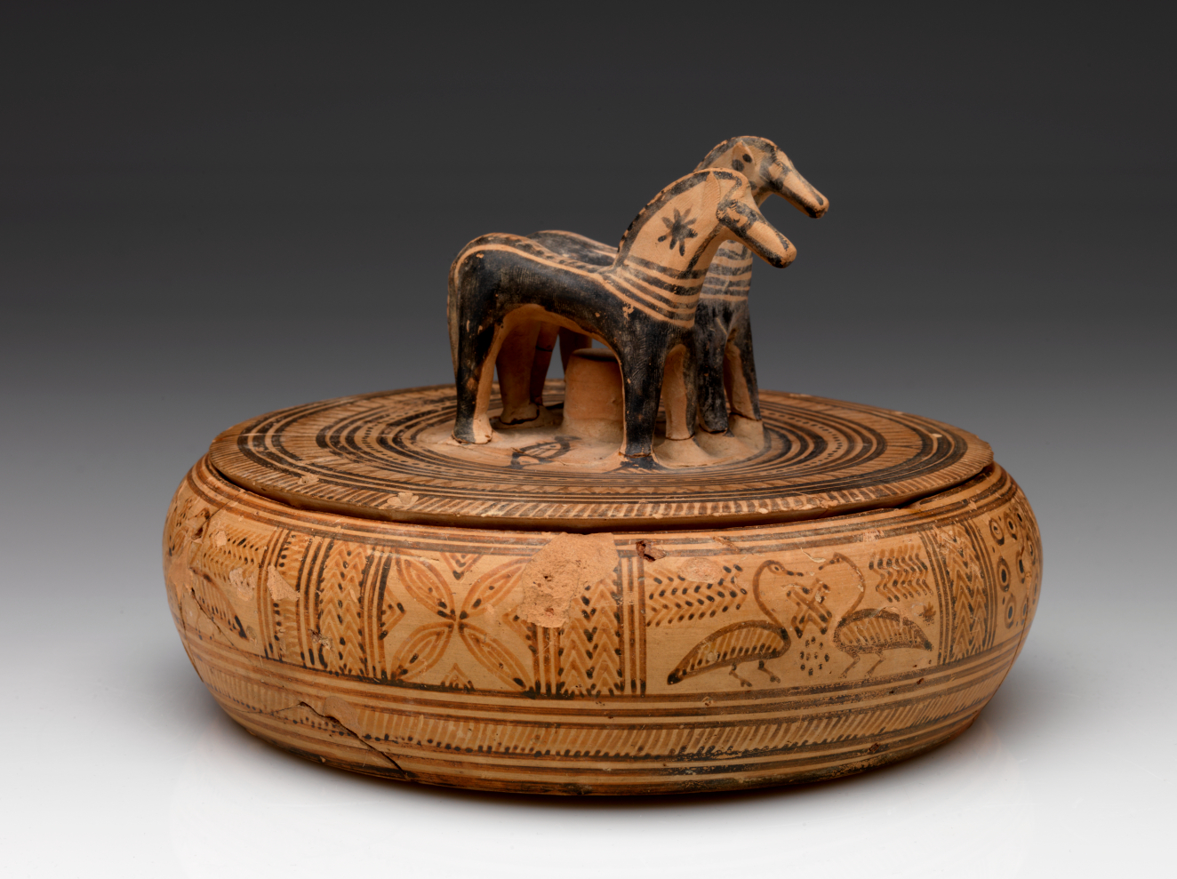 Side-view of a light-brown, short, wide, and round lidded box with a knob shaped as a pair of horses, decorated with patterns and natural imagery in black and red.