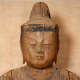 A close up of a weathered wooden Buddha’s face, with a serene expression and closed eyes. 