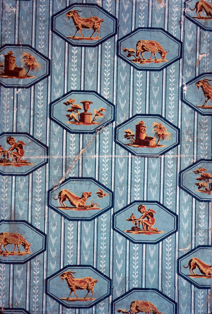 Vertically striped blue and white patterned wallcovering with repeating motifs of animals, figures, and buildings, in orange, within blue hexagonal frames overtop.