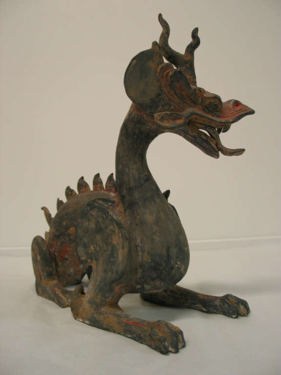 Quarter-view of an arched four-legged dragon with a lean body, open mouth, horned head, scaly back and a thin tail. The paint has worn into patches of browns and reds.