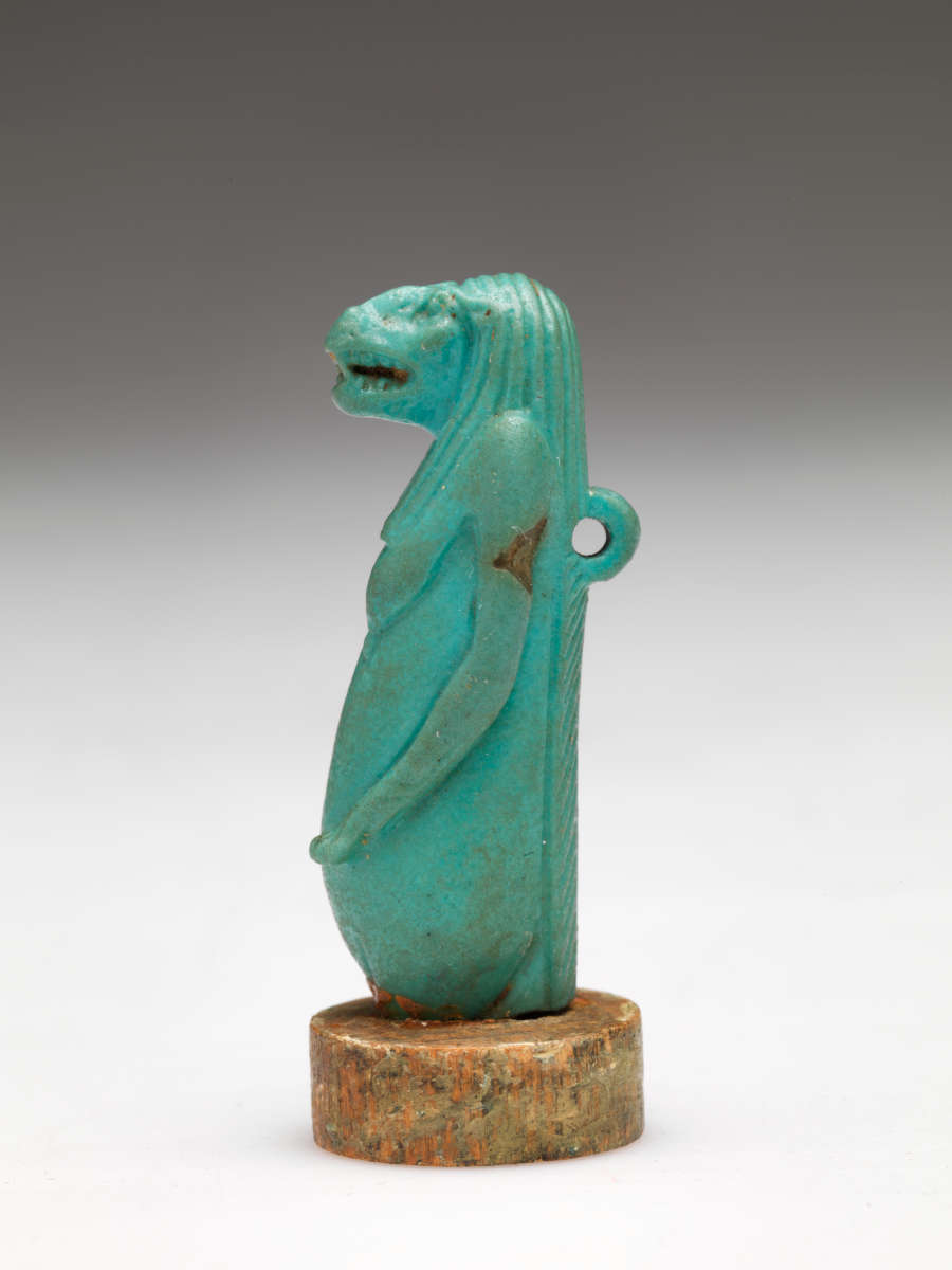 Side-view of the aquamarine sculpture of a nude anthropomorphic hippo-headed woman with bare breasts, long hair and arms, and sculpted loop on its back. It’s mounted on a stone disk.