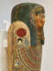 Angled view of a coffin with a gilded face, blue-black headdress and beard. A red sun above a falcon is painted on the shoulder and intricate patterns decorate the chest.