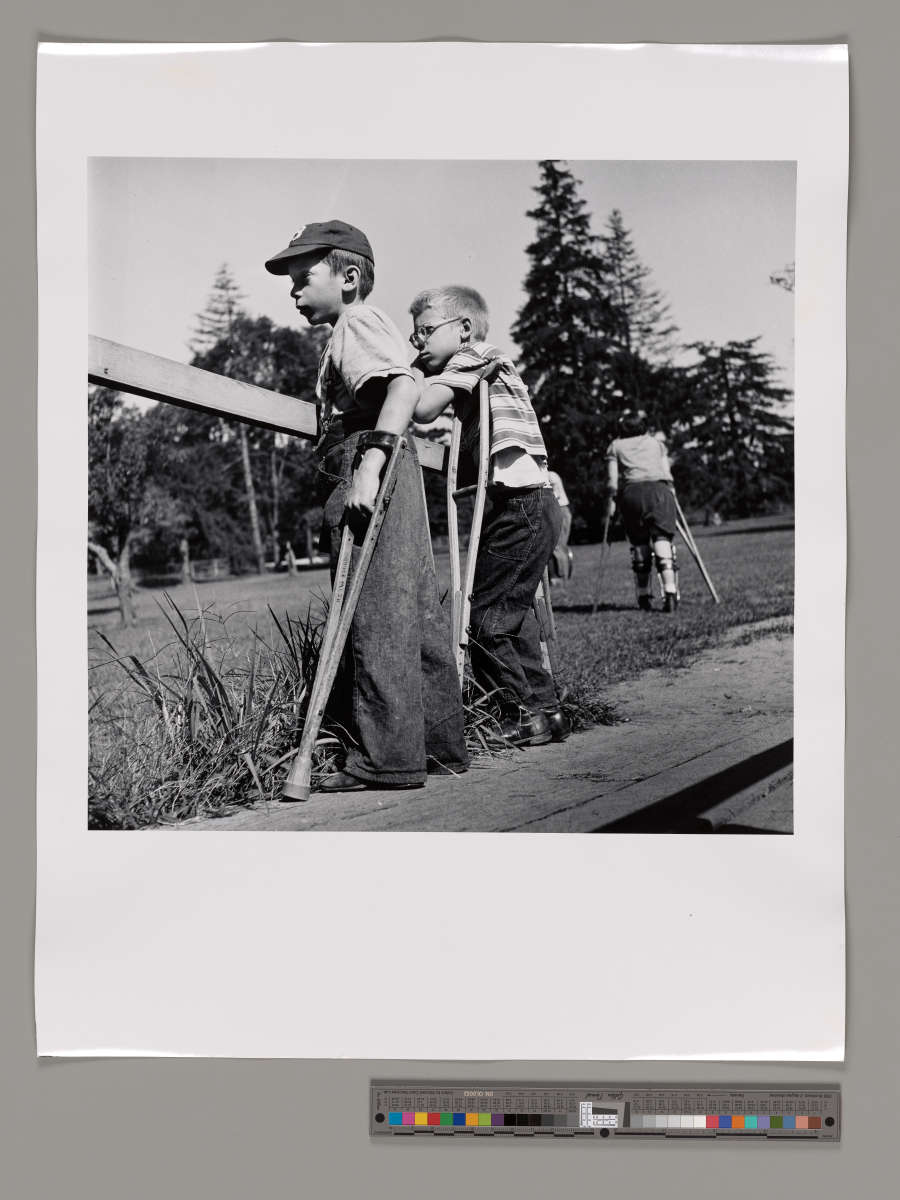 Grayscale photograph of two young boys standing with crutches and looking to the left. Another young child with crutches and leg braces walks into the park behind them.