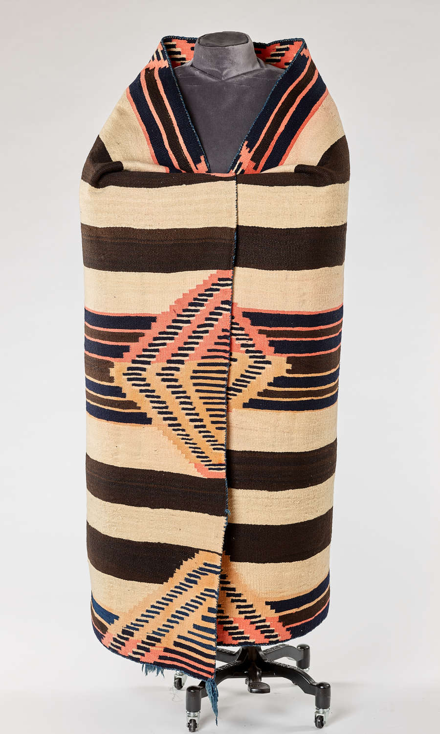 Black and cream striped woven blanket with salmon pink and yellow geometric patterns draped around a mannequin. The collar is symmetrical and the blanket end’s are overlapped and fastened.