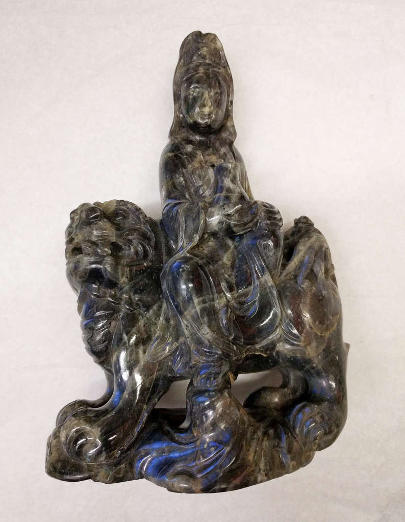Dark blue and brown-gray sculpture of a figure with a headdress seated on a four-legged creature with a mane. 