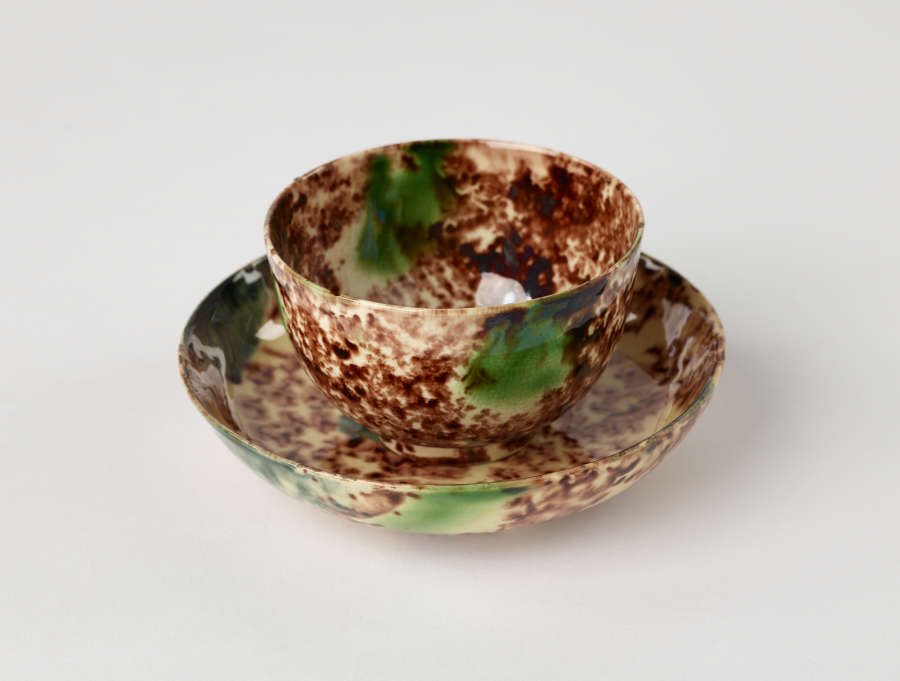  A handle-less cup and saucer that is white, brown, and green in color.