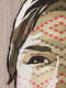 Close-up of the figure's right eye on a woven tapestry. Tiny red and beige diamond shapes with connecting thin lines form a geometric zig-zag pattern across the face.