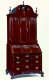 A tall standing cabinet made of polished mahogany wood with intricate metal handles on the lower drawers, fan-shaped carvings on the upper panels, and a rounded top featuring a curving inset.