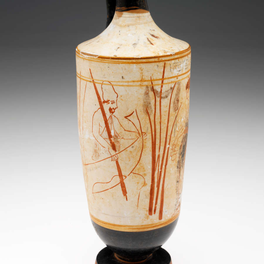 Close-up of the vase’s cream body. Visible are red illustrations of a man in a boat besides a cluster of reeds, holding a tall reed himself, bordered by golden stripes.