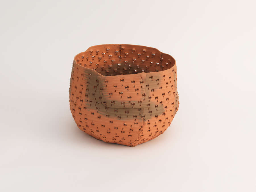 A small terracotta bowl with an uneven rim and a matte finish, the surface blending reddish and gray hues and punctured with tiny holes that form an irregular grid.