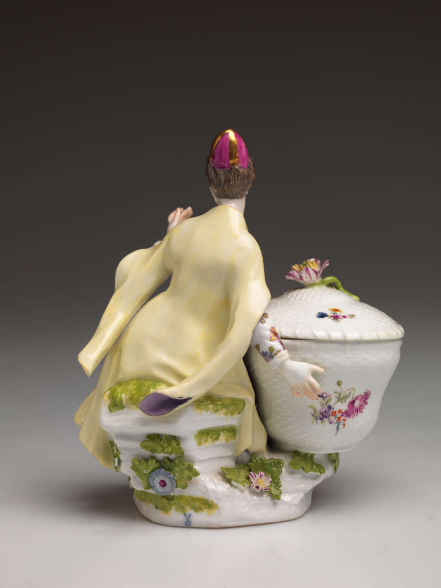 A sculptural figure sitting next to a large basket. The basket and figure are covered in floral decorations. The figure is wearing a hat, cream and purple clothing, and red shoes.