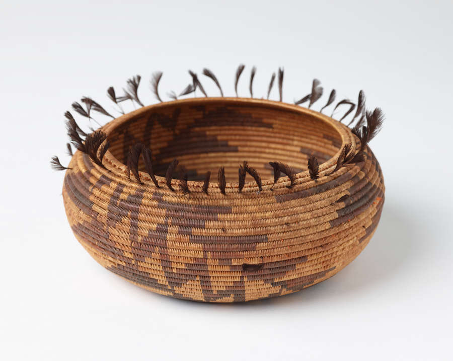 Woven tan and brown onion shaped bowl with geometric dark brown patterning and feather-like tassels emerging from the top edge of the bowl.