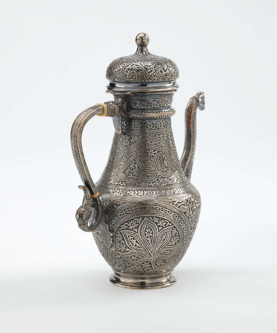 A silver coffee pot with detailed delicate decorations, handle with a lid hinged to it, and spout.