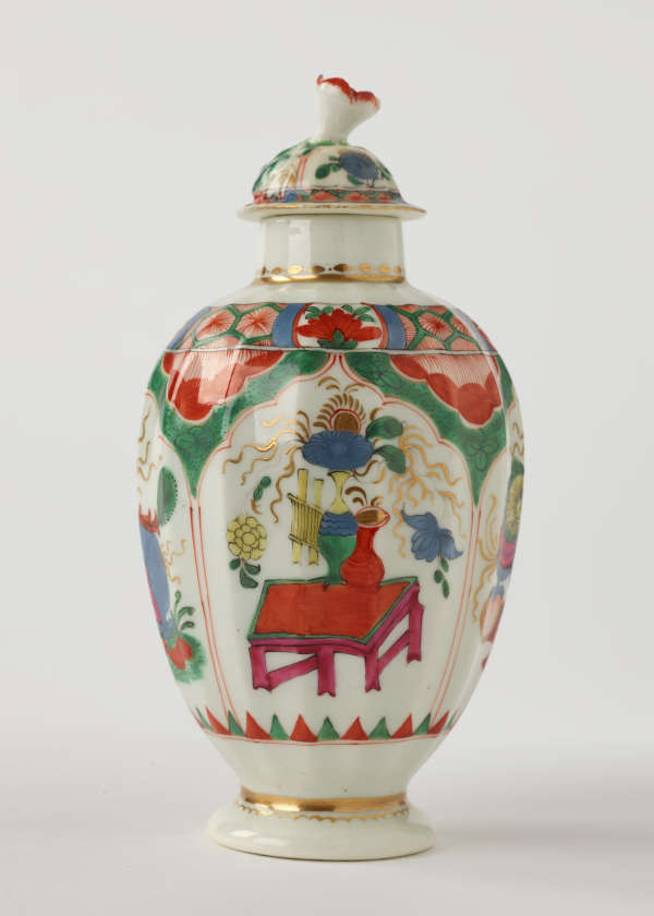 A tea caddy with a lid and green, red, blue, pink, yellow and gilded decorations. The finial is a sculptural flower white, red and green.