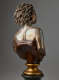 Back view of a bronze bust featuring hair in twists swept back and shoulder blades accentuated above a dark strapless top cut off diagonally and mounted on a gold base.