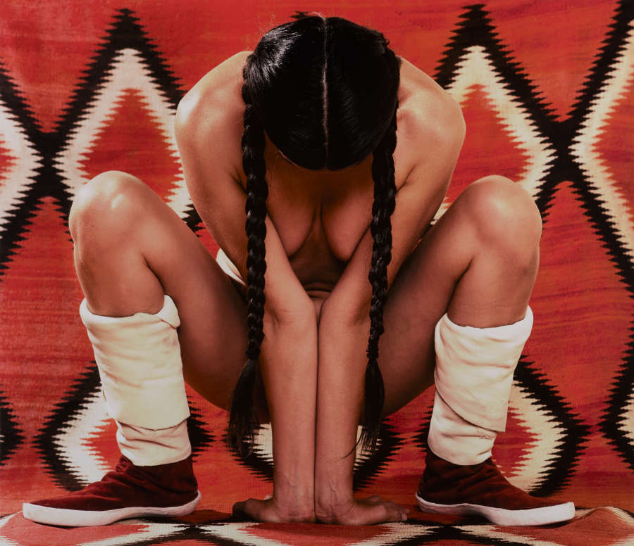 Nude person with braided hair and calf length shoes sits crouched, facing down, on a red geometric patterned backdrop. The person’s arms pressed together to cover their body.