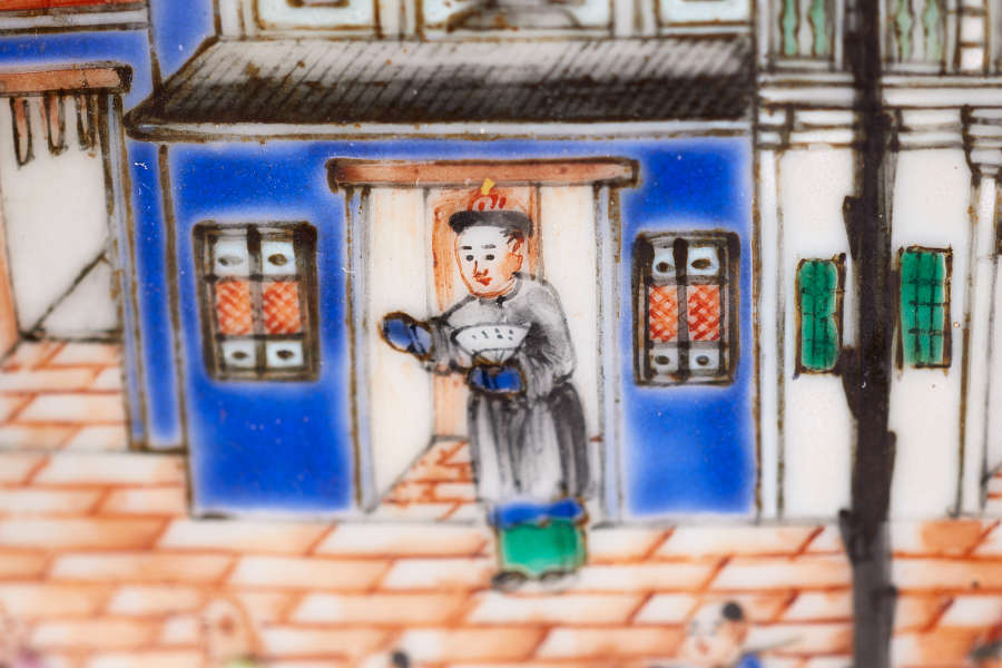 A close up view of a bowl with a painted figure, wearing a gray robe and holding a bowl in front of a doorway with blue walls and detailed windows.