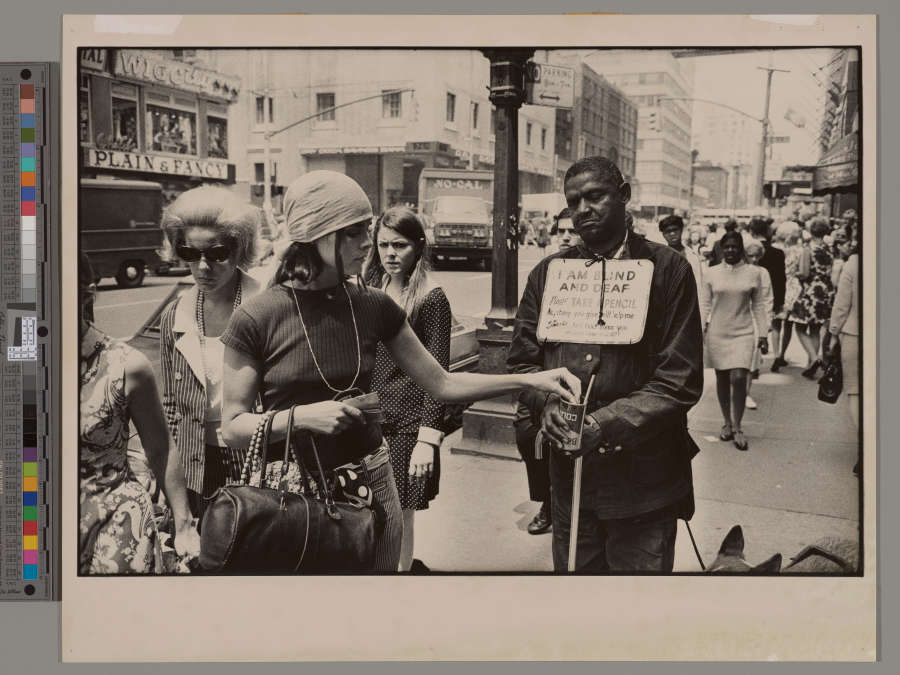 A grayscale photograph of a light-skinned person putting change into the cup of a dark-skinned person, who wears a sign around their neck that reads “I AM BLIND AND DEAF.”
