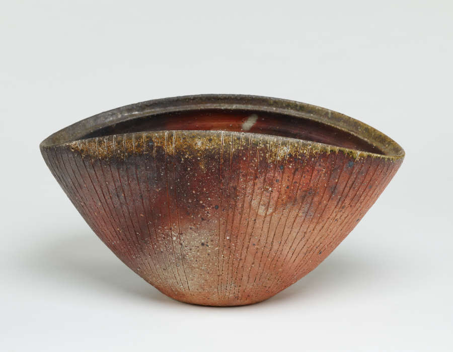 A deep clam shaped stone bowl with a wide opening and a gray-green rim. Shell like etchings outline the outer bowl which is dark brown on the left and a rich red brown on the right.