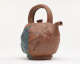 Hand-sculpted reddish-brown clay teapot with a wide arching handle and delicate spout. Along the left side, a rough blue-green patch of glaze is applied unevenly.