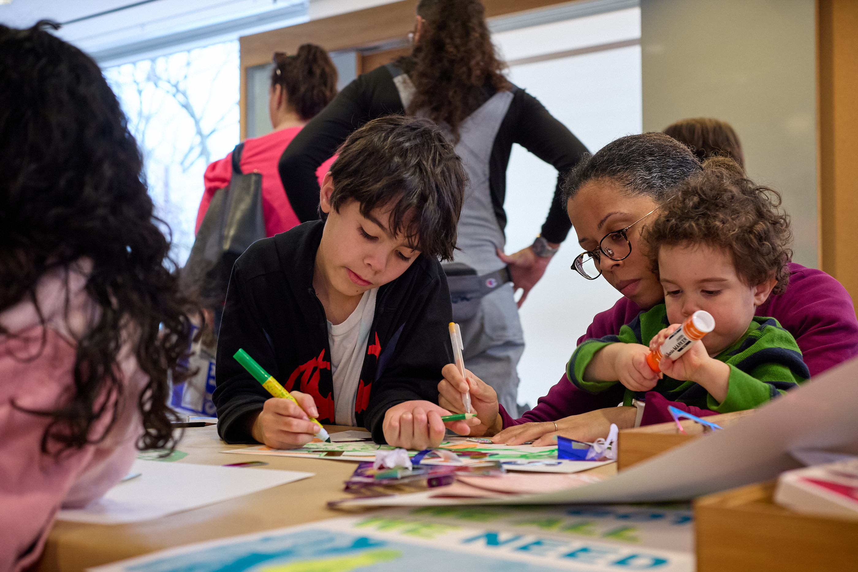 Children, assisted by adults, gather at a table, drawing and gluing with the array of markers and paper scraps spread across the table within a bright room.
