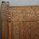 A close up view of a carved wooden panel featuring ornate calligraphy along the top and interwoven geometric star designs, framed by floral scrollwork.