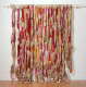 Textural installation featuring long bunches of beige yarn, tied in intervals with pink, red, and orange ribbons, hanging from a horizontal pole propped on a wall, creating a curtain-like display.