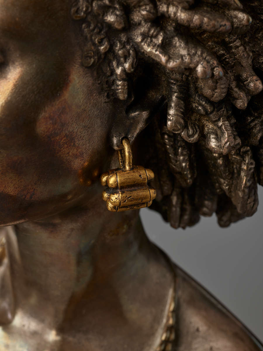 Close-up of left bronze cheek and ear displaying a chunky gold earring of clustered bullet-shaped forms dangling near dark bronze intricately coiled hair.