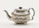 A silver teapot with a decorative handle, a rounded square body and spout with sculptural decorations.