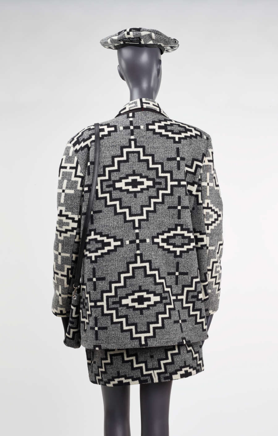 Back of a mannequin wearing a patterned, gray tweed blazer with a matching skirt, purse and hat. The matching patterns align creating a cohesive transition between garments. 