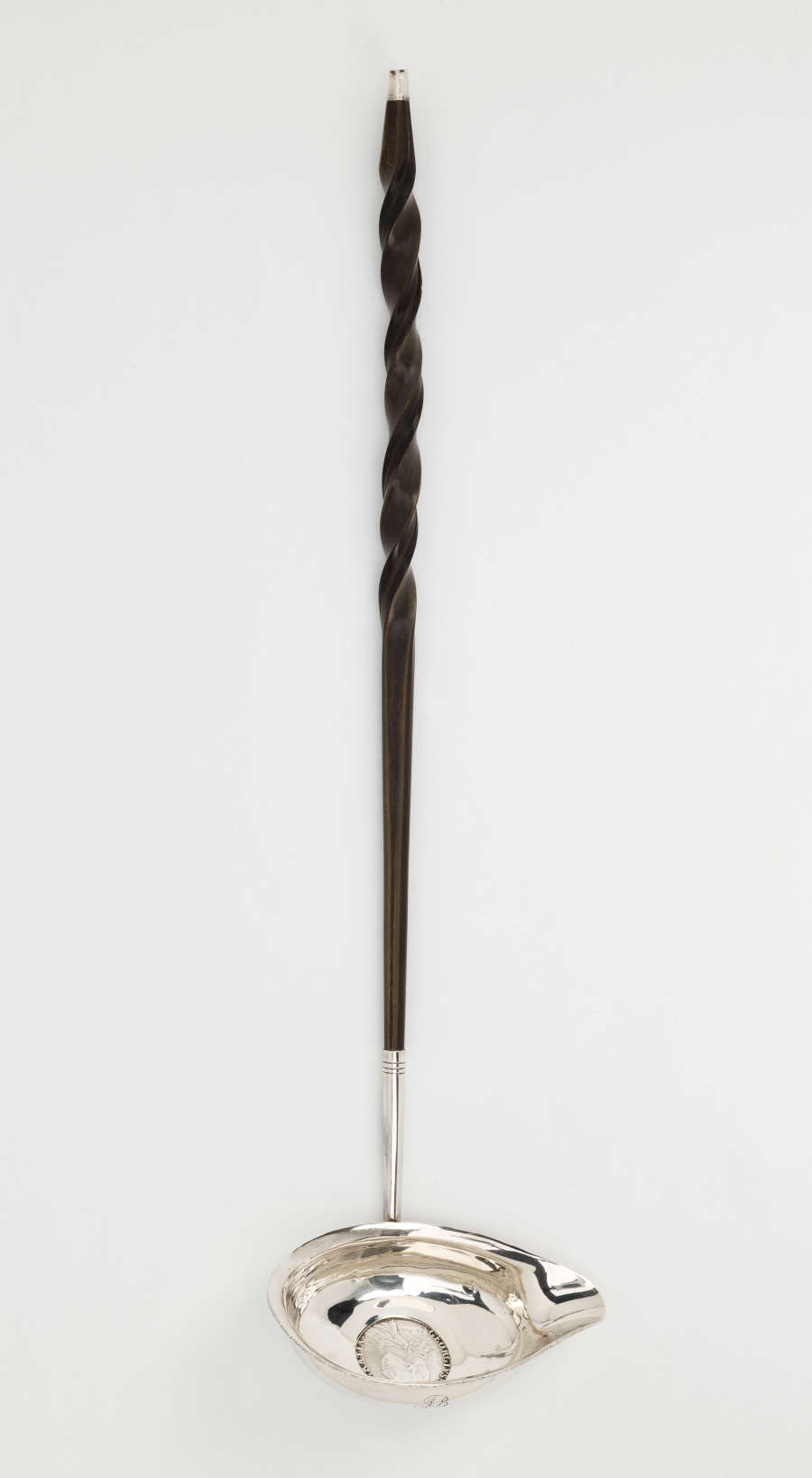 A silver and baleen coin ladle. The handle is twisted and dark and the ladle is silver with a small coin in the center.