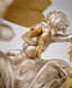 Close-up of the back view of a silver cherub with wavy hair, small, golden wings, draped sash over small chubby legs, and right arm extended. 