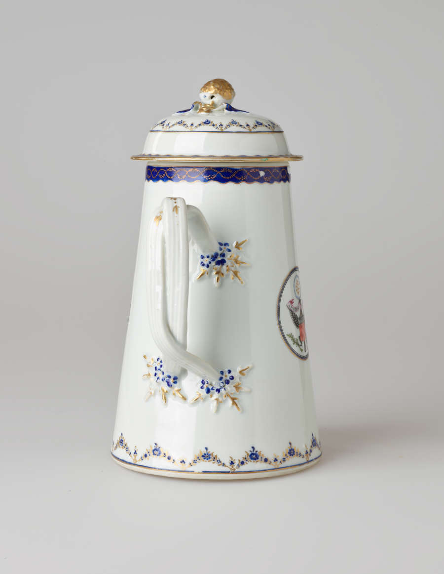 A white coffee pot with dark blue and gilded decorations, a straight spout, angular handle, and heraldry imagery in a circle on the body of the coffee pot.