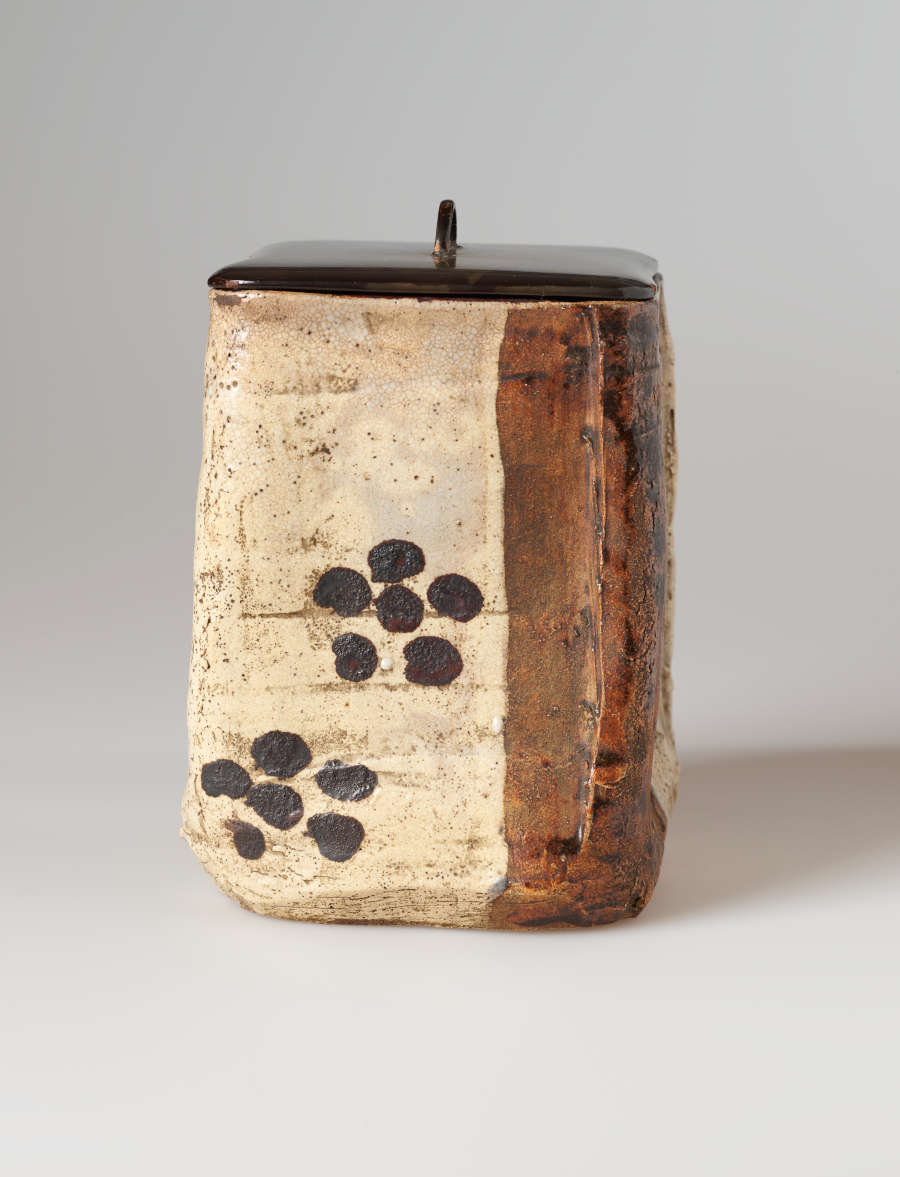 A textured, rectangular vessel. Its cream- and rust-colored surface is embellished with two six-dot designs. The flat dark lid is topped with a small knob.