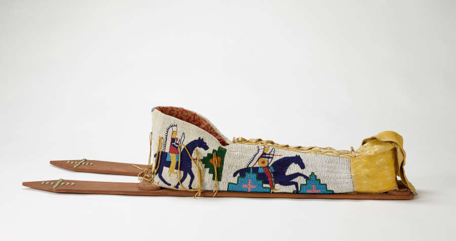 Beaded baby carrier lying flat on a surface. Back boards extend to the left. Beadwork designs in blue on white background depict horses, riders, and geometric decoration.
