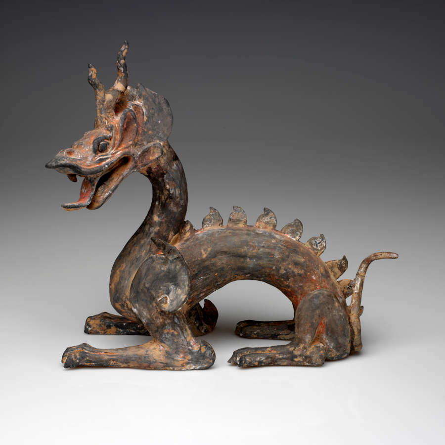 Side-view of an arched four-legged dragon with a lean body, open mouth, horned head, scaly back and a thin tail. The paint has worn into patches of browns and reds.