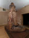 Side view of a large wooden Buddha statue, seated in meditation on a wooden platform in a warmly lit room, with visible cracks and repairs.