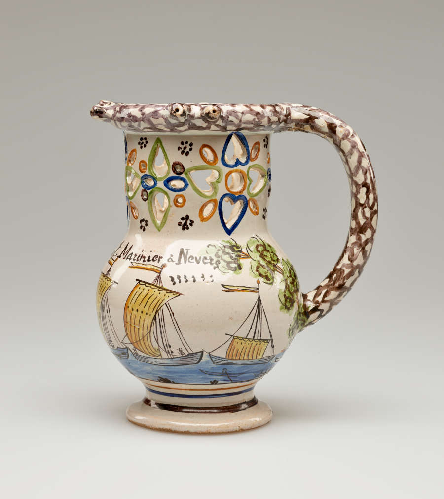 A jug with a rounded handle and pierced designs. The cream colored body is decorated with images of ships and a tree.
